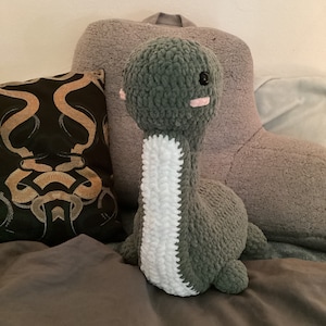 May include: A green crocheted dinosaur plush toy with a white stripe down its neck and pink cheeks. The toy is sitting on a gray and brown bedspread.