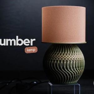 Puede incluir: Una lámpara de mesa moderna con una pantalla cilíndrica color melocotón y una base esférica verde con un diseño ondulado. La lámpara está etiquetada como "umber lamp" en texto blanco. La lámpara está sobre un soporte transparente.