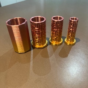 Puede incluir: Cuatro objetos cilíndricos metálicos con un degradado de oro a cobre. Los objetos están dispuestos en fila, el más alto a la izquierda. Tienen un diseño geométrico texturizado. El cilindro más alto mide unos 7 cm.