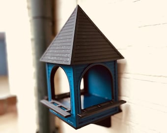 May include: A blue and black bird feeder with a peaked roof is mounted on a white brick wall. The bird feeder has a rectangular base with a small ledge and arched openings. The roof is dark gray and the body is a vibrant blue.