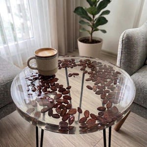 Può includere: Un tavolino in resina trasparente con chicchi di caffè marrone scuro incorporati nella superficie. Il tavolo ha gambe nere a forcina e una piccola tazza di caffè beige sopra. Una pianta da interno e una poltrona grigia sono sullo sfondo.