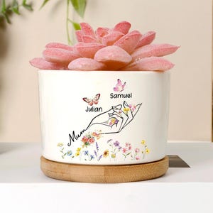 Könnte beinhalten: Ein weißer Keramik-Blumentopf mit einer rosa Sukkulente, auf einem Holzsockel. Der Topf hat ein handgezeichnetes Design mit Schmetterlingen und den Namen "Julian" und "Samuel". Das Wort "Mum" ist ebenfalls auf dem Topf geschrieben.
