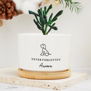 Könnte beinhalten: Ein weißer Keramiktopf mit einer kleinen grünen Sukkulente. Der Topf hat eine schwarze Linienzeichnung eines Hundes und den Text "NEVER FORGOTTEN Aurora". Der Topf steht auf einem Holzsockel.