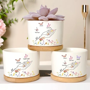 Könnte beinhalten: Drei weiße Keramik-Blumentöpfe mit Untersetzern aus Holz. Jeder Topf hat ein handgezeichnetes Design einer Hand, die Blumen, Schmetterlinge und das Wort "Mum" hält. Die Töpfe sind mit Namen personalisiert. Ein Topf enthält eine lila Sukkulente.