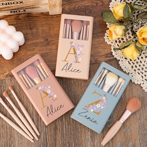 Könnte beinhalten: Drei Sets Make-up-Pinsel in rosa, blauen und goldenen Etuis mit Blumendesigns und personalisierten Namen. Die Etuis sind mit "Alice" und "Zenia" beschriftet.