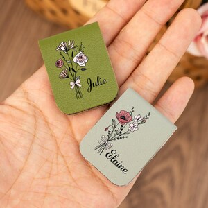 Könnte beinhalten: Zwei kleine, rechteckige Lesezeichen aus Leder. Eines ist olivgrün mit einem Blumenstrauß-Design und dem Namen "Julie". Das andere ist hellgrün mit einem Blumenstrauß-Design und dem Namen "Elaine".