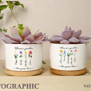 May include: Two white ceramic plant pots with artificial succulents, each on a wooden coaster. The pots have the text "Mama's garden" and "love grows here" with floral and name details. The succulents are purple and green.