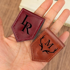 Könnte beinhalten: Zwei magnetische Lesezeichen aus Leder. Eines ist weinrot mit den Initialen "LR" in Schwarz. Das andere ist braun mit dem Buchstaben "M" und einem Hirschgeweih-Design in Schwarz. Beide sind genäht.