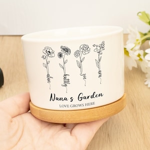Può includere: Un vaso di ceramica bianco con una base in bambù. Il vaso ha un contorno nero di quattro fiori con nomi scritti sui gambi: Ava, Zoe, Kaitlyn e Alan. Il testo "Nana's Garden" e "Love Grows Here" è stampato sotto i fiori.