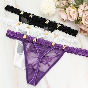 Könnte beinhalten: Drei String-Tangas in Schwarz, Weiß und Lila, jeweils mit dem Wort "BABY" in goldenen Buchstaben auf dem Bund. Die Unterwäsche hat Spitzendetails und ein zartes Design. Blumige Akzente sind im Hintergrund zu sehen.