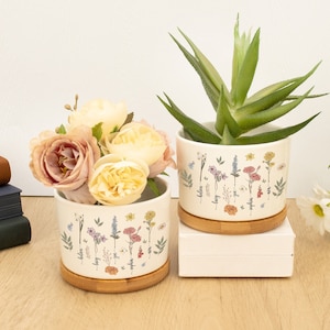 May include: Two white ceramic flower pots with a bamboo base. The pots feature a floral design with the names of different flowers written on them. One pot contains a succulent plant and the other has a bouquet of pink and yellow flowers.
