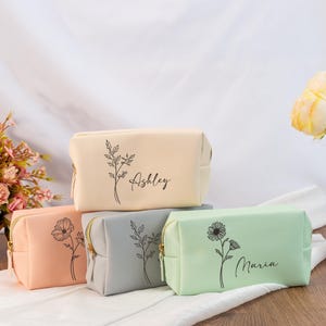 Peut inclure: Quatre trousses de maquillage aux couleurs pastel : beige, pêche, gris et vert menthe. Chaque trousse présente un motif floral noir et un nom en écriture cursive. Les trousses ont des fermetures éclair dorées.