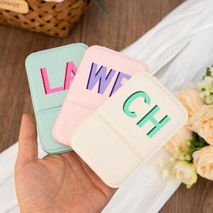May include: Three pastel-coloured pill cases are held in a hand. Each case has a different colour and features a two-letter word in a contrasting colour: LA, WE, and CH. The cases are rectangular with rounded corners.