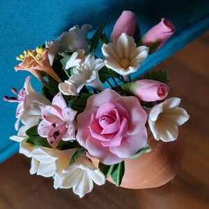 Puede incluir: Una pequeña maceta de terracota llena de un ramo de flores artificiales. Las flores están hechas de tela rosa, blanca y naranja y están dispuestas de forma realista. El ramo incluye rosas, lirios y otros tipos de flores.