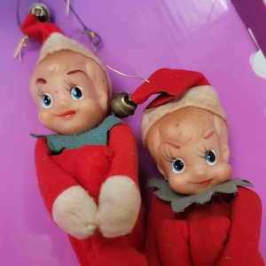 Puede incluir: Dos adornos de elfo de Navidad vintage con gorros rojos y blancos, cuellos verdes y cuerpos de fieltro rojo. Los elfos tienen caras pintadas con ojos azules y están sosteniendo sus manos juntas.