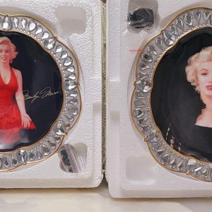 May include: Two decorative plates featuring images of Marilyn Monroe. Each plate has a black background with a portrait of Monroe and her signature. The plates are framed with a silver-colored, scalloped edge adorned with clear, faceted stones.