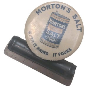 May include: Vintage Morton's Salt tin lid and scraper. The lid is round, cream-colored, and features the Morton's Salt logo with the text "When it rains it pours." The scraper is rectangular with a dark handle.