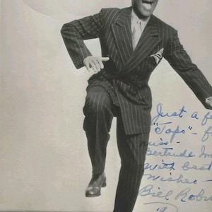 May include: Black and white vintage photograph of a man in a pinstripe suit and bowler hat, mid-dance. The image includes handwritten text and a signature in the lower left corner. The man is smiling.