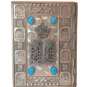 May include: A silver-colored, rectangular book cover with intricate embossed designs. Features a crown, two tablets, and six turquoise-colored stones. The cover is adorned with detailed illustrations and Hebrew text.
