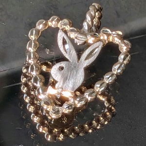 Puede incluir: Un colgante plateado con el logotipo del conejito Playboy dentro de un marco en forma de corazón. El corazón está delineado con pequeñas cuentas facetadas, creando un diseño texturizado y llamativo. El colgante es probablemente de metal.