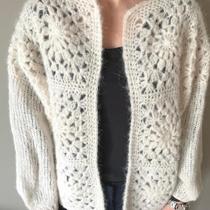 Può includere: Un cardigan bianco lavorato a uncinetto con un motivo traforato. Il cardigan ha una vestibilità ampia e maniche lunghe.