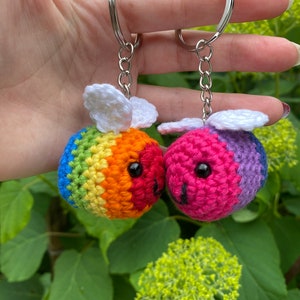 Può includere: Due portachiavi a forma di ape all'uncinetto con schemi di colori arcobaleno e viola. Le api hanno ali bianche e occhi neri. Sono attaccati a portachiavi argentati.
