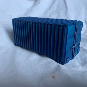 Könnte beinhalten: Ein blaues Modell eines Schiffscontainers mit wellenförmigen Metallwänden und einer geschlossenen Tür.