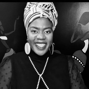 May include: A woman wearing a black mock neck shirt with a pearl necklace and large oval earrings. She is wearing a black and white patterned head wrap.