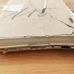 Puede incluir: Un libro desgastado, cubierto de cart&oacute;n marr&oacute;n, con lomo blanco y algunas p&aacute;ginas visibles. El libro est&aacute; ligeramente da&ntilde;ado y tiene algunas roturas en la cubierta.