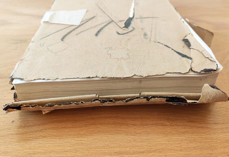Puede incluir: Un libro desgastado, cubierto de cart&oacute;n marr&oacute;n, con lomo blanco y algunas p&aacute;ginas visibles. El libro est&aacute; ligeramente da&ntilde;ado y tiene algunas roturas en la cubierta.