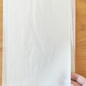Puede incluir: Dos hojas de papel pergamino blanco, apiladas una encima de la otra, sobre una superficie de madera.
