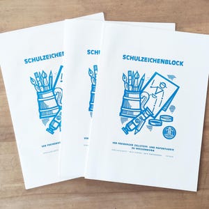 以下が含まれることがあります： 青いイラストが描かれた、3冊の白いSCHULZEICHENBLOCKノート。ノートには、筆や鉛筆がいっぱい入ったマグカップ、絵の具のチューブ、人物の絵が描かれています。