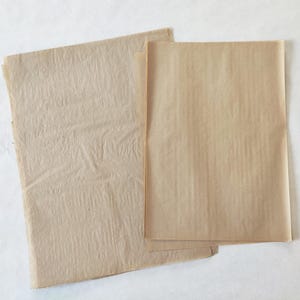 Könnte beinhalten: Zwei rechteckige Bögen hellbraunes Papier. Ein Bogen auf der linken Seite hat eine faltige, strukturierte Oberfläche, während der Bogen auf der rechten Seite glatt und flach ist. Beide Papierbögen sind dünn, durchscheinend und naturfarben.
