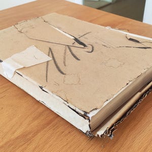 Puede incluir: Una caja de cart&oacute;n desgastada con una etiqueta blanca y marcas negras. La caja est&aacute; ligeramente da&ntilde;ada y tiene un aspecto desgastado y envejecido.