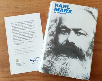 Puede incluir: Una biografía de Karl Marx, titulada "Karl Marx Biographie", con un retrato de Marx en la portada. Una tarjeta adjunta con texto en alemán y una firma está al lado del libro. El libro y la tarjeta están sobre una superficie de madera.
