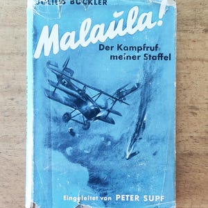 Könnte beinhalten: Vintage Buchcover mit dem Titel "Malaula!" in weißer Schrift. Der blaue Hintergrund zeigt eine Illustration von Flugzeugen. Der Text "Der Kampfruf meiner Staffel" ist ebenfalls sichtbar. Der Autor, Julius Buckler, ist ebenfalls sichtbar.