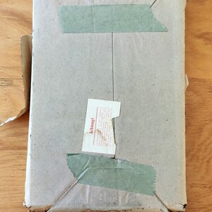 Puede incluir: Una caja de cart&oacute;n marr&oacute;n con cinta adhesiva verde y una etiqueta blanca que dice "Achtung!" en letras rojas.