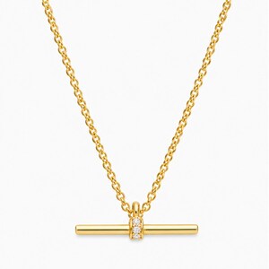 May include: A gold necklace featuring a horizontal bar pendant with three small, clear stones. The chain is a fine link design. The necklace is set against a plain white background.