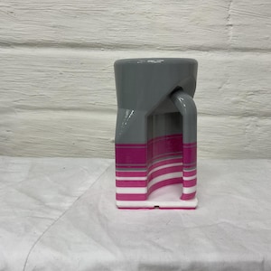 May include: A gray and pink ceramic mug with a unique geometric design. The mug features a gray top and handle, with pink and white stripes on the body. The mug is approximately 4 inches tall.