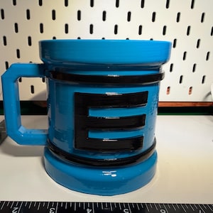 Puede incluir: Una taza azul brillante con una gran letra negra "E" en la parte delantera. La taza tiene forma cilíndrica con un asa y bandas negras alrededor de la parte superior, media e inferior. La taza está sobre una superficie blanca.