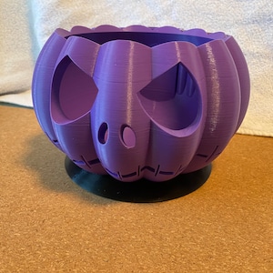 May include: A purple 3D printed pumpkin planter with a carved face. The planter has a black base.