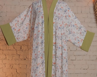 Könnte beinhalten: Weißer Bademantel mit floralem Muster und grünen Zierleisten und Manschetten. Der Bademantel hat ein zartes Muster aus orangefarbenen, blauen und violetten Blumen. Die Robe wird auf einer Schaufensterpuppe vor einer Backsteinmauer präsentiert.