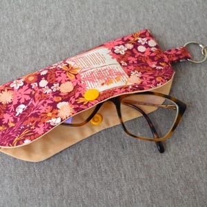 Könnte beinhalten: Brillenetui mit floralem Muster in Rosa-, Orange- und Weißtönen auf bordeauxfarbenem Hintergrund. Es hat einen Druckknopfverschluss und einen silberfarbenen Schlüsselanhänger. Im Etui befindet sich eine Schildpattbrille.