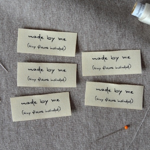 Könnte beinhalten: Fünf beige Stoffetiketten mit dem Text "made by me (tiny flaws included)" in schwarzer Schrift. Zwei Nadeln mit blauen und orangenen Köpfen sind sichtbar, zusammen mit einer weißen Garnrolle und einer Nadel.