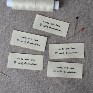 Könnte beinhalten: Mehrere rechteckige Stoffetiketten mit dem handgeschriebenen Text "made with love & wild frustration". Eine cremefarbene Garnrolle, Stecknadeln und eine Nadel deuten auf ein Näh- oder Bastelprojekt hin.