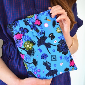 Könnte beinhalten: Eine blaue Clutch mit einem skurrilen Alice im Wunderland-Druck. Der Stoff zeigt schwarze Silhouetten von Alice, Blumen und anderen ikonischen Bildern. Die Clutch hat einen Klappenverschluss und wird von einer Person gehalten.
