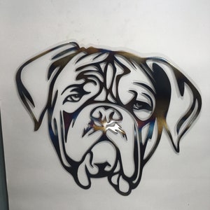 May include: Metal wall art of a dog's face. The artwork is cut from metal and has a dark colour with hints of blue and gold. The dog's features are stylized, with large, floppy ears and a prominent snout. Suitable for home decor.