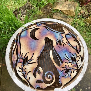 May include: A circular metal fire pit with a cut-out design of two stylized animals and foliage. The metal has a blue, purple, and orange iridescent finish.