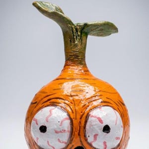Könnte beinhalten: Eine handbemalte Keramikskulptur einer runden, karottenartigen Figur mit einem grünen Stiel oben. Die Figur hat zwei große, hervorstehende weiße Augen mit roten Adern, einen kleinen schwarzen Mund und dunkelblaue Füße an der Basis des Körpers.