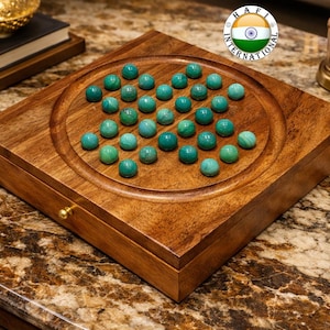May include: A wooden solitaire game board with a circular recessed playing area. The board is filled with 33 teal-colored marbles. The board is made of a rich, dark wood and has a small drawer. A gold-colored knob is on the drawer.